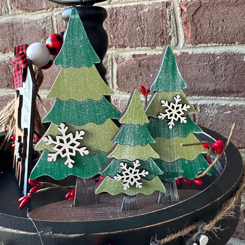 May include: Three wooden Christmas trees with a distressed white paint finish. The trees are decorated with white snowflakes and are arranged on a black tiered tray.