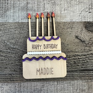 May include: Wooden birthday cake with purple frosting and five candles. The cake is decorated with white dots and the words "Happy Birthday" and "Maddie".