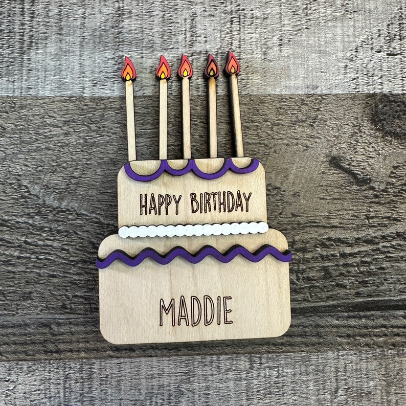 May include: Wooden birthday cake with purple frosting and five candles. The cake is decorated with white dots and the words "Happy Birthday" and "Maddie".