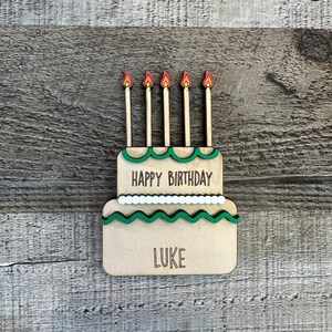 May include: Wooden birthday cake with green frosting and five candles. The cake says "Happy Birthday" and "Luke".