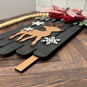 Puede incluir: Un letrero de madera negro con un reno marr&oacute;n y copos de nieve blancos. El letrero est&aacute; decorado con un lazo rojo y blanco a rayas y una campana.