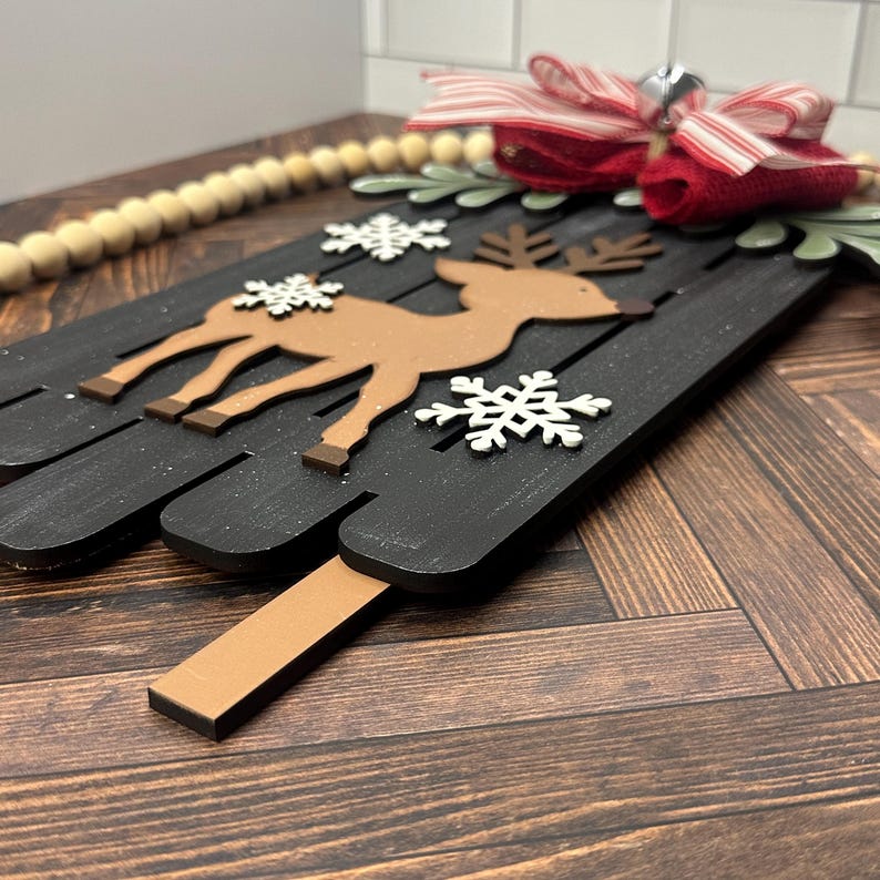 Puede incluir: Un letrero de madera negro con un reno marr&oacute;n y copos de nieve blancos. El letrero est&aacute; decorado con un lazo rojo y blanco a rayas y una campana.