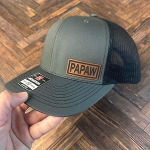 May include: A dark green and black trucker hat with a brown leather patch that says "PApaw" on the front.
