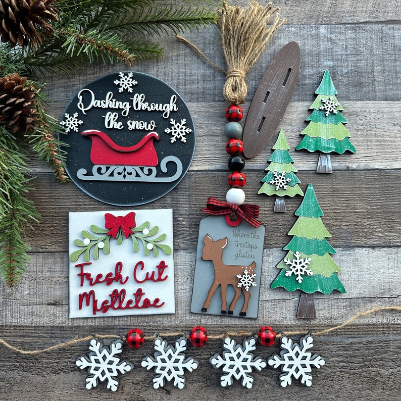 May include: A set of Christmas-themed wooden signs with various sayings, including "Dashing through the snow" with a red sleigh, "Fresh Cut Mistletoe" with a red bow, and "Where the treetops glisten" with a deer. The signs are decorated with snowflakes and red and black buffalo plaid.