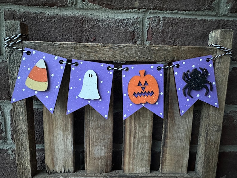 May include: Purple and white Halloween banner with a ghost, pumpkin, spider, and candy corn. The banner is strung on a wooden dowel.