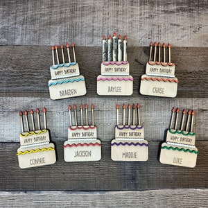 May include: Eight wooden birthday cake decorations with candles and names. The cakes are decorated with different colors and patterns. The names are Braeden, Raylee, Chase, Connie, Jackson, Maddie, Luke.