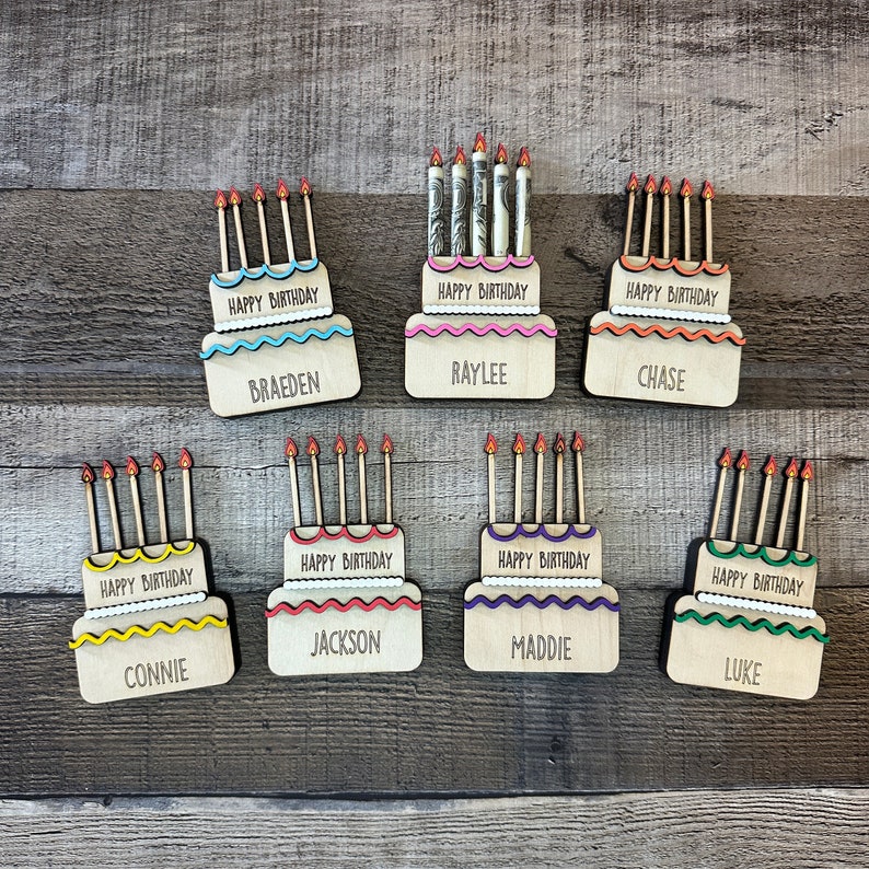 May include: Eight wooden birthday cake decorations with candles and names. The cakes are decorated with different colors and patterns. The names are Braeden, Raylee, Chase, Connie, Jackson, Maddie, Luke.