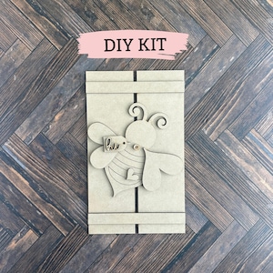 Op de afbeelding: DIY-knutselset met een houten uitsnijding van een bij met het woord "hi" op een label. De bij heeft zwarte en gele strepen en een hart op zijn lichaam. De set bevat alle benodigde materialen om een decoratief stuk te maken.