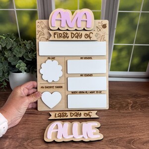 Puede incluir: Un letrero de madera del primer y último día de clases con el nombre "AVA" en letras rosas. El letrero incluye espacios para escribir detalles sobre el año escolar. También se incluye un letrero separado con el nombre "ALLIE".