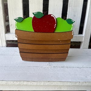Puede incluir: Una cesta de madera llena de tres manzanas. Las manzanas est&aacute;n pintadas de rojo y verde. La cesta es marr&oacute;n con un acabado moteado blanco.