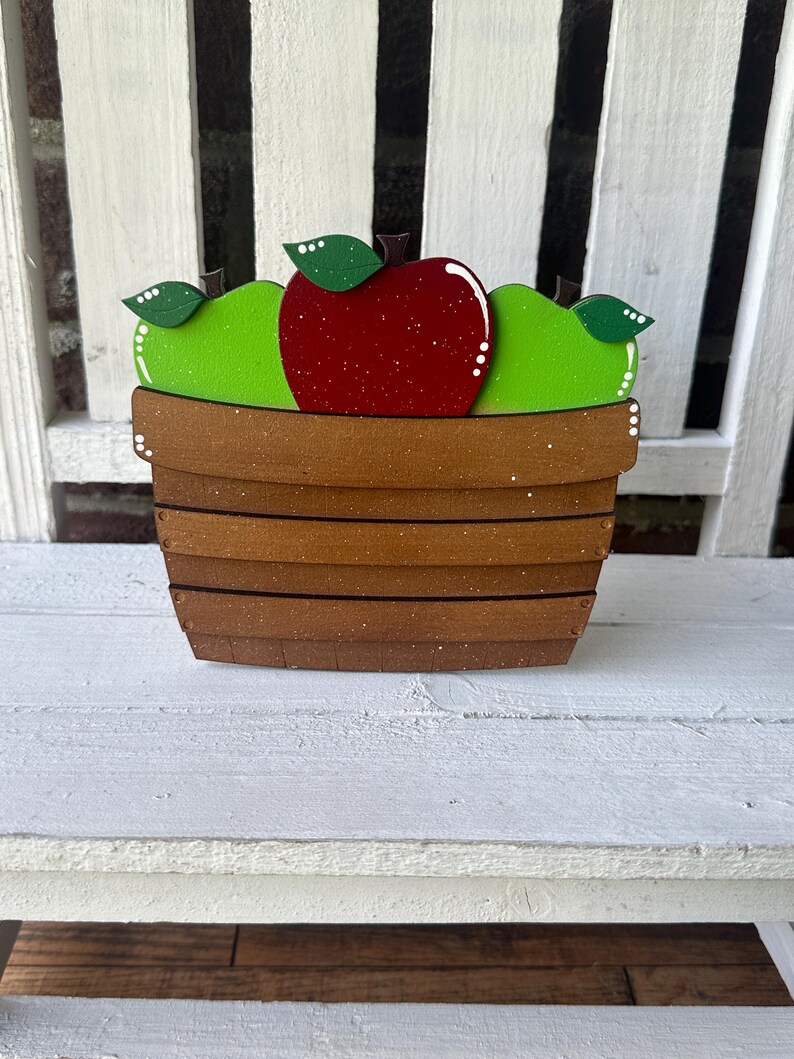 Puede incluir: Una cesta de madera llena de tres manzanas. Las manzanas est&aacute;n pintadas de rojo y verde. La cesta es marr&oacute;n con un acabado moteado blanco.