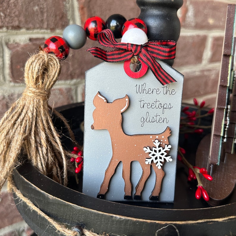 May include: A wooden Christmas tag with a white background and a brown deer silhouette. The tag has the text "Where the treetops glisten" written on it. The tag is decorated with a red and white buffalo plaid ribbon and a white snowflake.