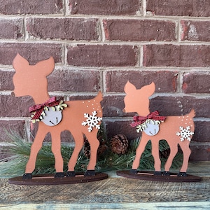 May include: Two wooden reindeer decorations, one larger than the other, with red and white checked bows and bells. The reindeer are standing on a brown wooden base.
