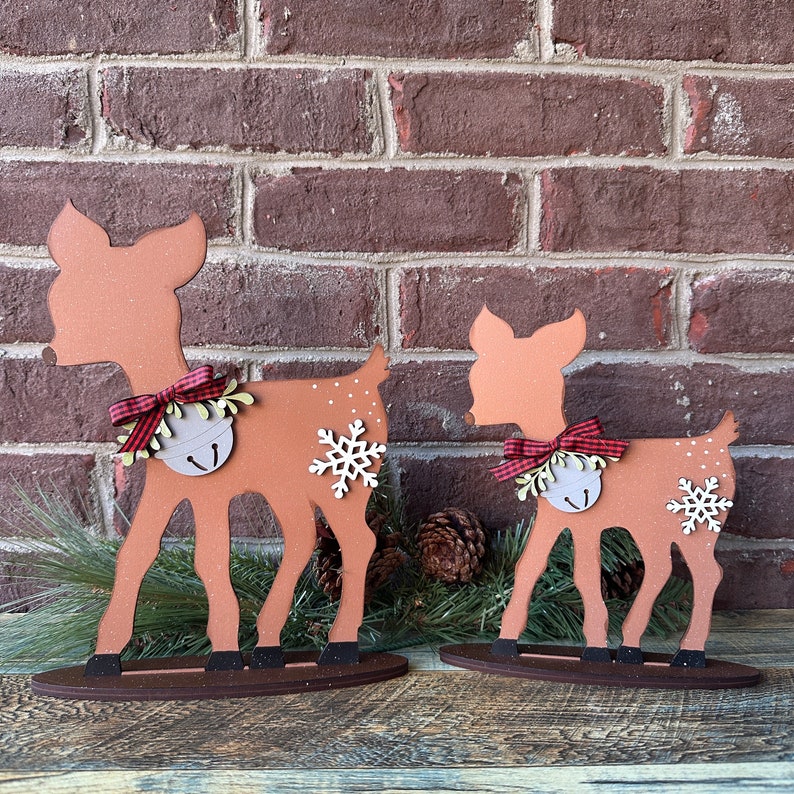 May include: Two wooden reindeer decorations, one larger than the other, with red and white checked bows and bells. The reindeer are standing on a brown wooden base.