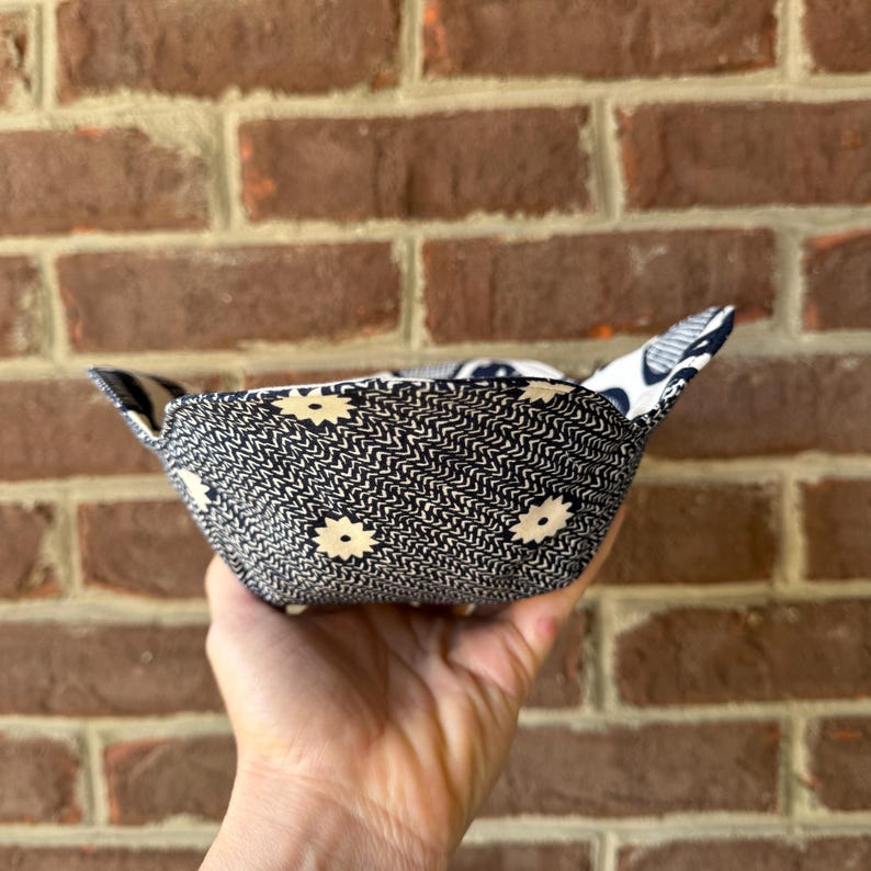 May include: A handmade fabric bowl with a navy blue and cream floral pattern. The bowl is square with folded corners and is held in a hand. The background is a brick wall.