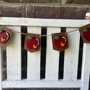 Puede incluir: Una pancarta de madera con forma de manzana roja con hojas verdes y corazones amarillos cuelga de una valla blanca de madera. La pancarta est&aacute; unida con un cordel.