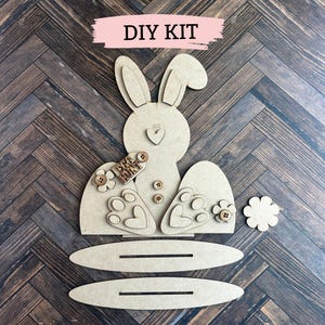 May include: DIY craft kit featuring a bunny rabbit with a "Egg Hunt" sign, two oval-shaped bases, a flower, and small wooden embellishments.