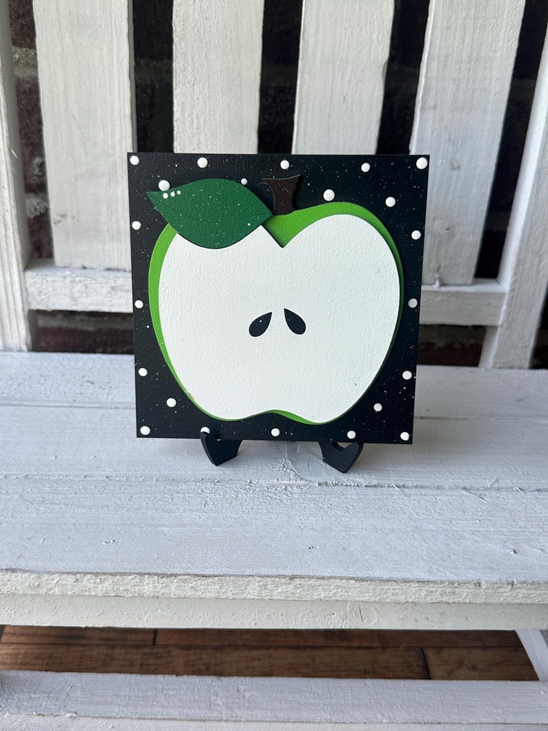 Puede incluir: Una manzana verde con un centro blanco y un tallo negro est&aacute; pintada sobre un fondo negro con lunares blancos. La manzana est&aacute; sobre un peque&ntilde;o caballete de madera.