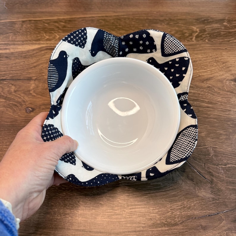 May include: A white ceramic bowl sits in a navy blue and cream-colored bowl cozy. The cozy features a bird pattern. The bowl is held by a person's hand. The background is a wooden surface.