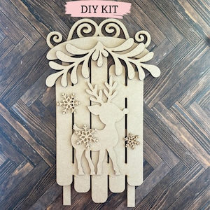 Puede incluir: Kit de bricolaje para una decoraci&oacute;n de trineo de reno navide&ntilde;o de madera. El kit incluye un reno, un trineo, copos de nieve y una guirnalda de hojas.