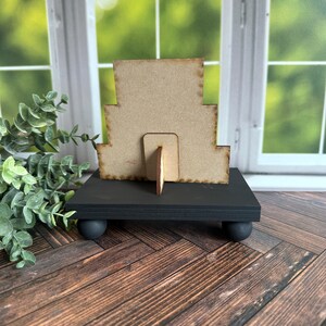May include: A tiered wooden picture frame with a black stand and round feet. The frame is made of unfinished wood and has a rustic look.