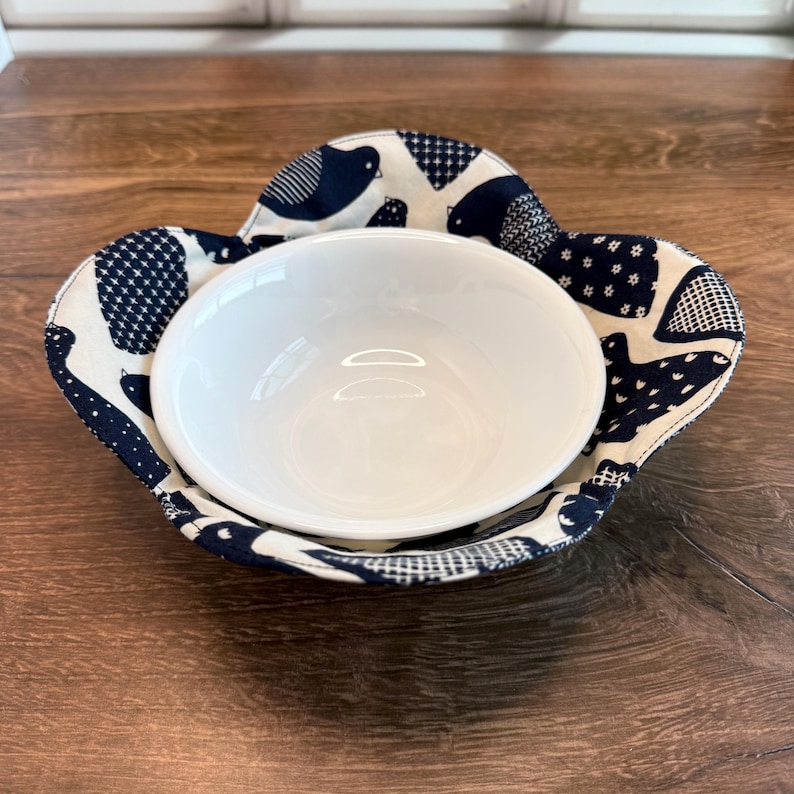 May include: A white ceramic bowl nested in a navy blue and cream fabric bowl cozy. The fabric features a pattern of birds and geometric shapes. The cozy is on a dark wood surface.