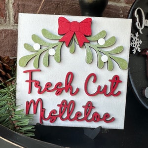 May include: A white wooden sign with a red bow and green mistletoe sprigs. The sign says "Fresh Cut Mistletoe" in red glitter letters.
