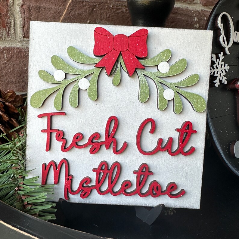 May include: A white wooden sign with a red bow and green mistletoe sprigs. The sign says "Fresh Cut Mistletoe" in red glitter letters.