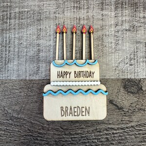 May include: A wooden birthday cake with blue and white frosting and five candles. The cake says "Happy Birthday" and "Braeden".
