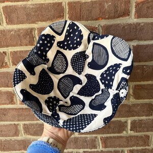 May include: A white and navy blue patterned bucket hat. The hat features a repeating pattern of stylized birds in various designs. The hat is made of fabric and has a rounded crown and a wide brim. The hat is held up by a person.