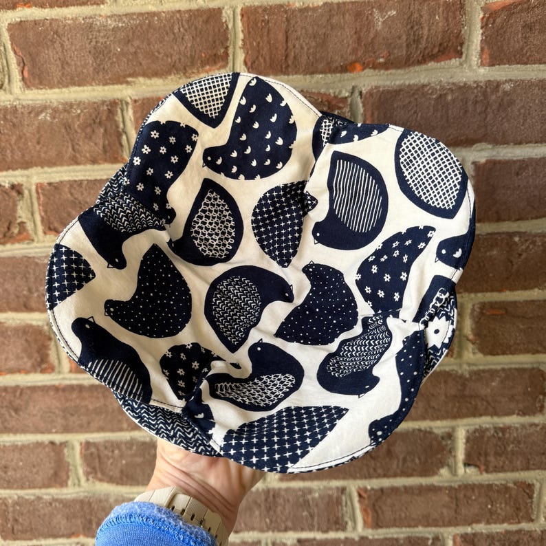 May include: A white and navy blue patterned bucket hat. The hat features a repeating pattern of stylized birds in various designs. The hat is made of fabric and has a rounded crown and a wide brim. The hat is held up by a person.