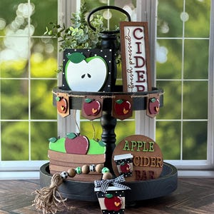 Puede incluir: Una bandeja de dos niveles de madera negra decorada con letreros y adornos de madera con tem&aacute;tica oto&ntilde;al. El nivel superior presenta un letrero que dice "Cidra reci&eacute;n prensada" y una manzana de madera con una hoja verde. El nivel inferior presenta un letrero que dice "Bar de sidra de manzana" y una cesta de manzanas de madera con una manzana roja y hojas verdes. La bandeja est&aacute; decorada con una guirnalda de cuentas de madera y una borla de arpillera.