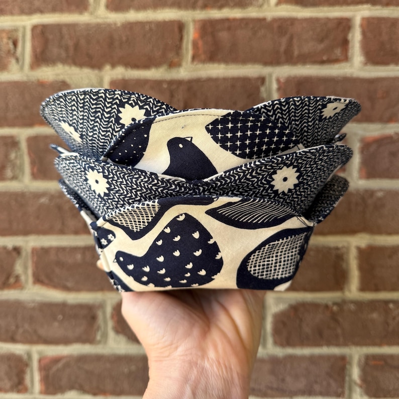 May include: A stack of four fabric bowls with a navy blue and cream bird and geometric pattern. The bowls are held in a hand and have a scalloped edge. The bowls are made of fabric and are likely used for serving food.