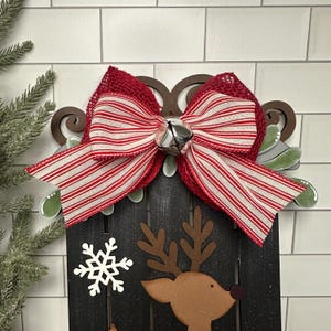 Puede incluir: Un trineo de Navidad de madera negra con un lazo a rayas rojas y blancas, una campana plateada y un recorte de reno. El trineo est&aacute; decorado con un copo de nieve blanco.