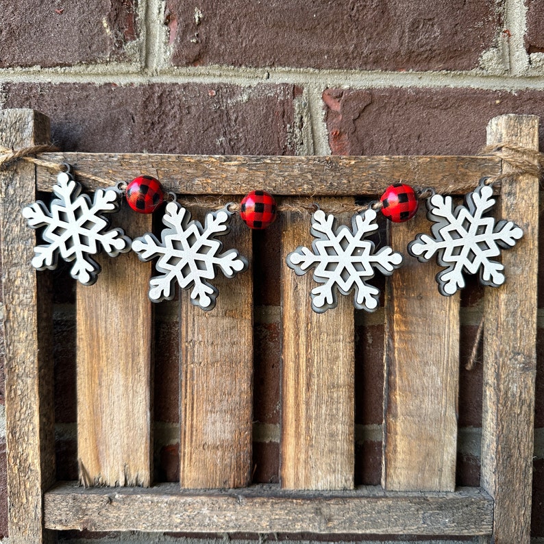 May include: A white wooden snowflake garland with red and black buffalo plaid ornaments. The garland is hanging on a wooden slat wall.
