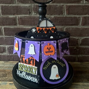May include: A black tiered stand with a purple base and white polka dots. The stand features Halloween-themed decorations, including a ghost, a pumpkin, a spider, and a candy corn. The top tier has a black car with a white ghost and the words "BOO" on a banner. The bottom tier has three signs that read "EEK! SPOOKY" and "Halloween". There is also a round sign with a ghost and the words "trick or treat".
