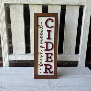Puede incluir: Un letrero de madera con marco marr&oacute;n y fondo blanco. El letrero dice "fresh pressed CIDER" en letras rojas con purpurina dorada.