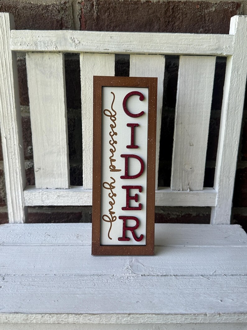 Puede incluir: Un letrero de madera con marco marr&oacute;n y fondo blanco. El letrero dice "fresh pressed CIDER" en letras rojas con purpurina dorada.