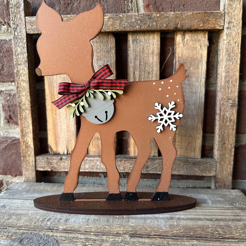 May include: A wooden cutout of a brown reindeer with a red and white checked bow, a bell, and a snowflake. The reindeer is standing on a brown base.