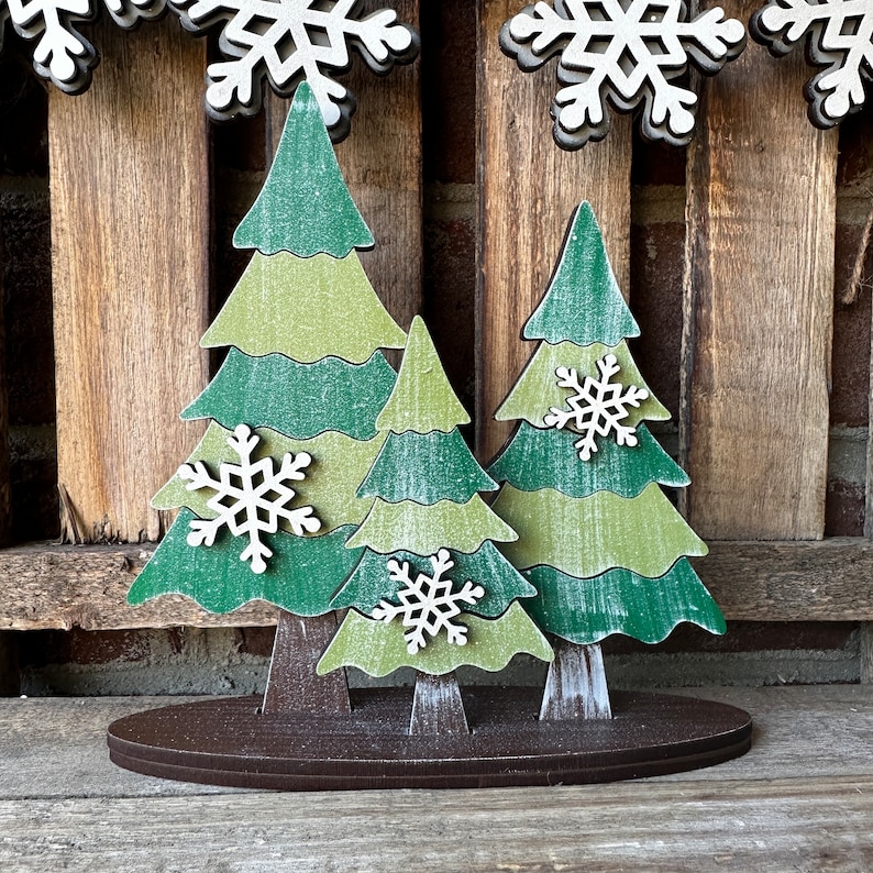 May include: A wooden Christmas tree decoration with three green trees with white snowflakes on each tree. The trees are standing on a brown base.