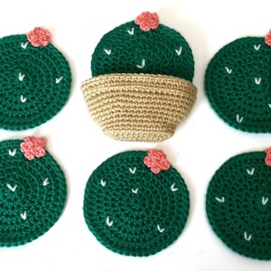 May include: Handmade crochet cactus coasters in a set of six. Each coaster is a dark green circle with white stitch details and a small pink flower. A beige crochet basket is also included.