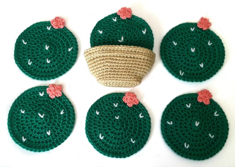 May include: Handmade crochet cactus coasters in a set of six. Each coaster is a dark green circle with white stitch details and a small pink flower. A beige crochet basket is also included.