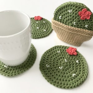 May include: A white ceramic mug rests on a green crocheted cactus coaster. Additional cactus coasters are visible, featuring a green body, white accents, and a small pink flower. One coaster is in a tan crocheted pot. The coasters are handmade.