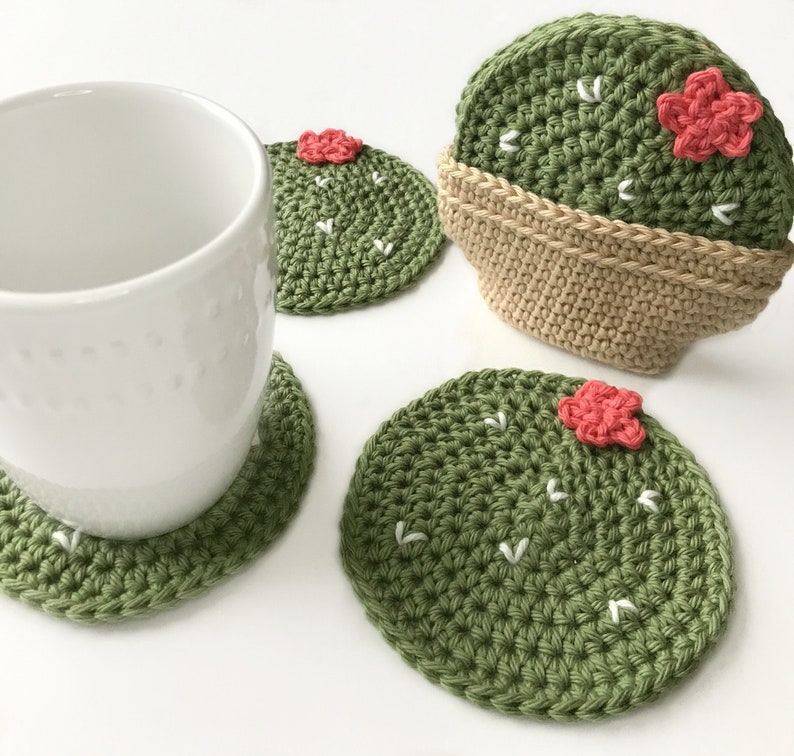 May include: A white ceramic mug rests on a green crocheted cactus coaster. Additional cactus coasters are visible, featuring a green body, white accents, and a small pink flower. One coaster is in a tan crocheted pot. The coasters are handmade.