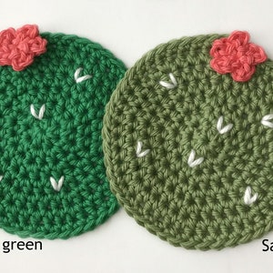 May include: Two crocheted cactus-shaped coasters. One is bright green, the other sage green. Each has a small, coral-colored flower and white stitched details. The text "Bright green" and "Sage" are below the coasters.