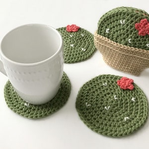 May include: A white ceramic mug rests on a green crocheted cactus coaster. Additional cactus coasters are visible, featuring a green body, white accents, and a pink flower. One coaster is in a tan crocheted pot. The coasters are handmade.