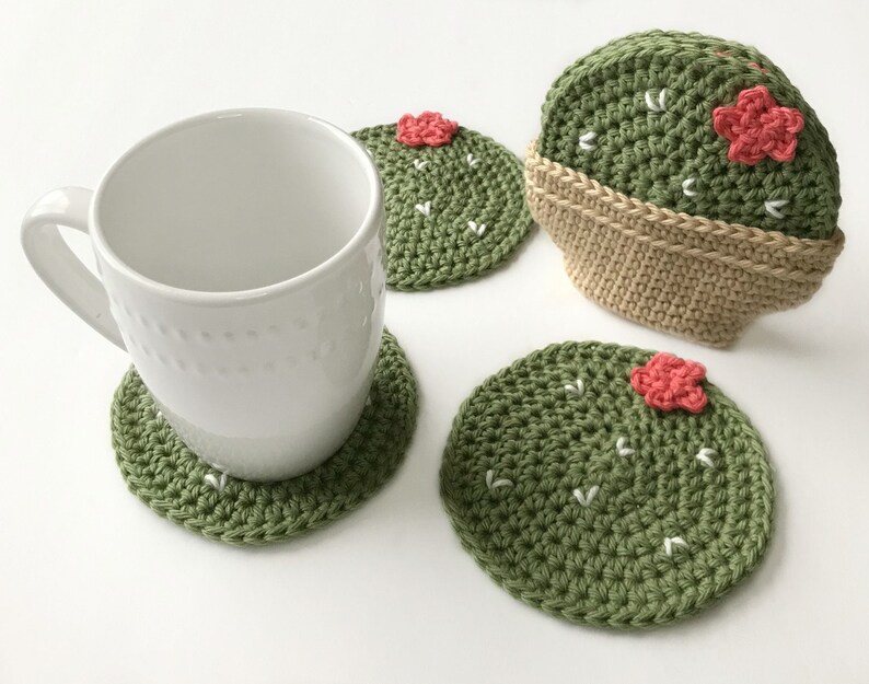 May include: A white ceramic mug rests on a green crocheted cactus coaster. Additional cactus coasters are visible, featuring a green body, white accents, and a pink flower. One coaster is in a tan crocheted pot. The coasters are handmade.