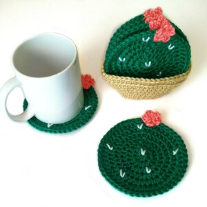 May include: Handmade crochet cactus coasters in green with white accents and small pink flowers. One coaster is under a white mug, and two others are displayed, one in a woven basket. The coasters are round and add a decorative touch.