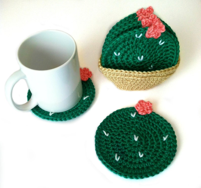 May include: Handmade crochet cactus coasters in green with white accents and small pink flowers. One coaster is under a white mug, and two others are displayed, one in a woven basket. The coasters are round and add a decorative touch.