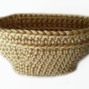 May include: A hand-crocheted basket in a light tan color. The basket has a rounded shape with a wide opening and a textured surface. The stitches are clearly visible, creating a detailed pattern. This item is suitable for home decor.